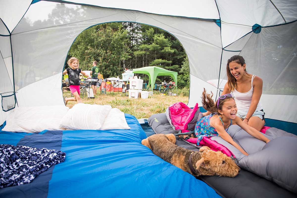 Happy Mother with Daughter jumping into the Eureka! Space Camp Family Camping Tent landing on sleeping bag right next to pet dog