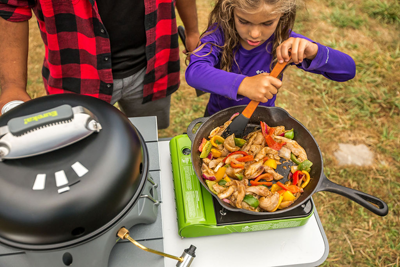 A young girl stirs a pan full of stir-fry on a camp stove, while her data watches over her shoulder. 