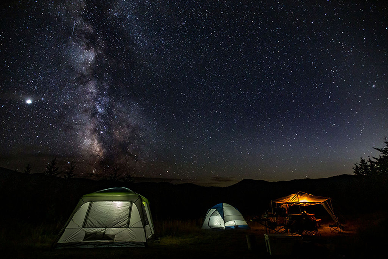 Two tents and a shelter illuminated by light at night, the Milky Way visible in the starry night sky. 