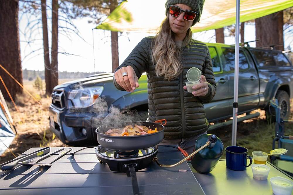 Woman sprinkling salt and pepper into a meaty pepper dish cooked on the Jetboil HalfGen camp stove on Eureka!'s Camp Kitchen Table while car camping