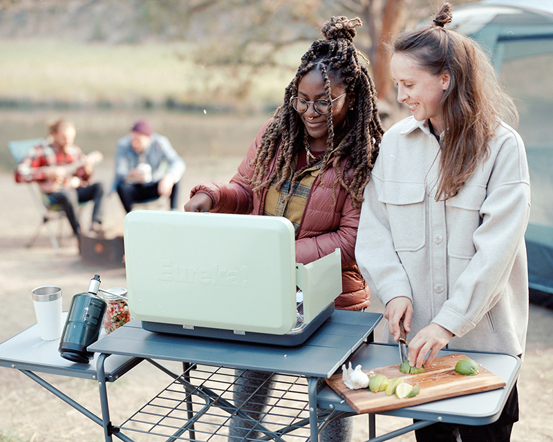 Two young women cook on a green Eureka Camp stove at a campsite. 