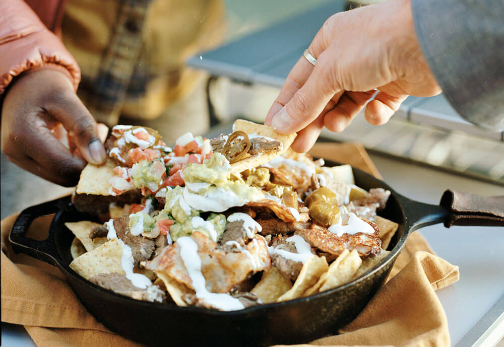 Hands reaching to grab carne asada nachos from a cast iron skillet