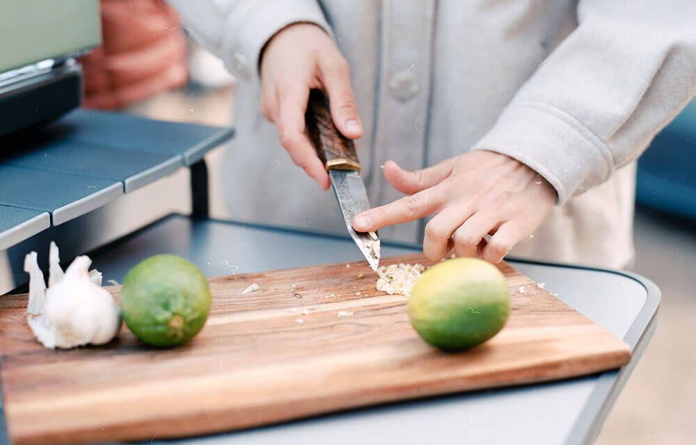 Finger sliding off garlic from a cutting knife on a wooden chopping board with garlic and limes sitting on the Eureka! Camp Kitchen table