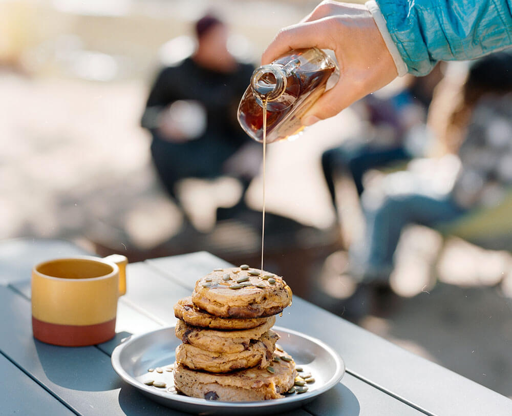 Pouring syrup from jar onto stack of platted chocolate chip pumpkin pancakes sitting on Eureka! Camp Kitchen