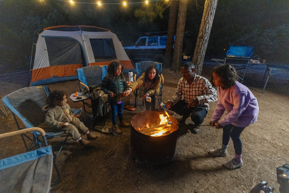 Family at campsite gathered around camp fire roasting marshmallows on stick 