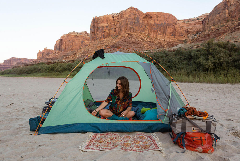 Woman sitting in her Kohana camp tent on a beach with rocky mountains in the background