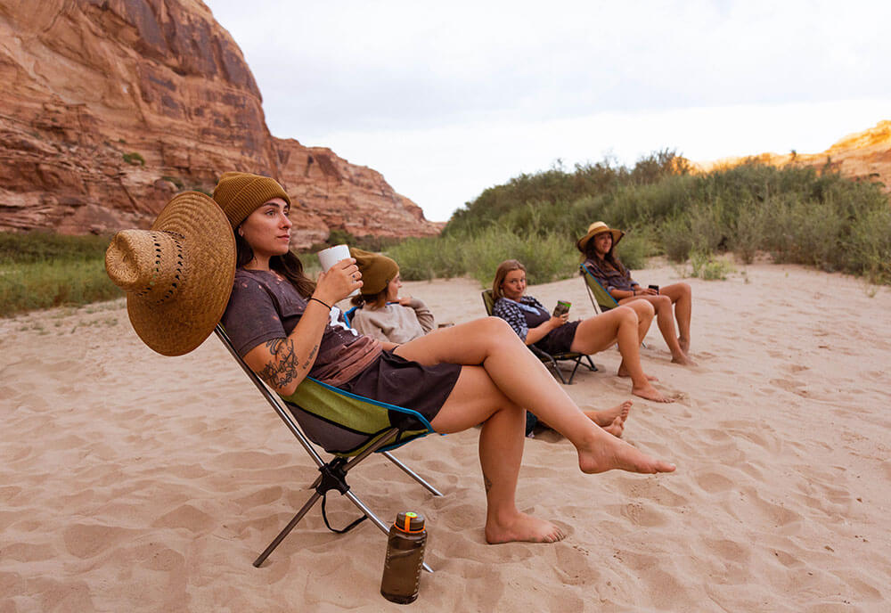 Group of 4 female friends relaxing at sandy campsite with beverages in hand while sitting in Eureka! Tagalong camp chairs