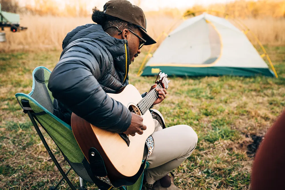 Caziah Franklin sits in a camp chair and strums an acoustic guitar with a tent pitched in the background. 