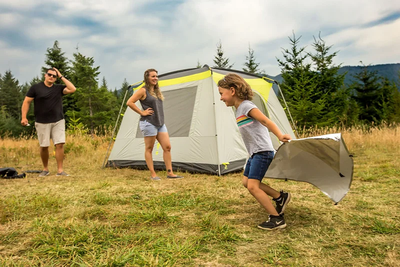 A young child runs around the campsite laughing, holding a tent fly behind him like a cape.