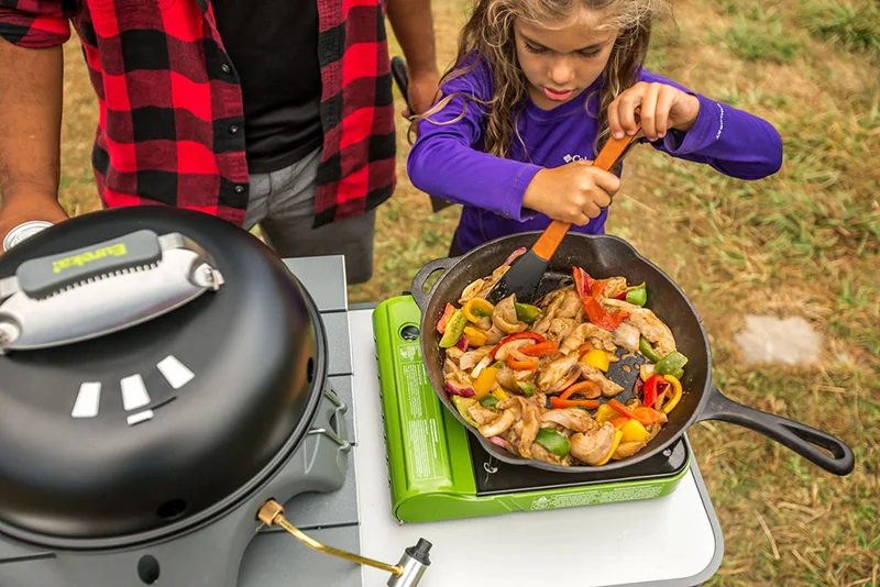 A young girl stirs a pan full of stir-fry on a camp stove, while her data watches over her shoulder. 